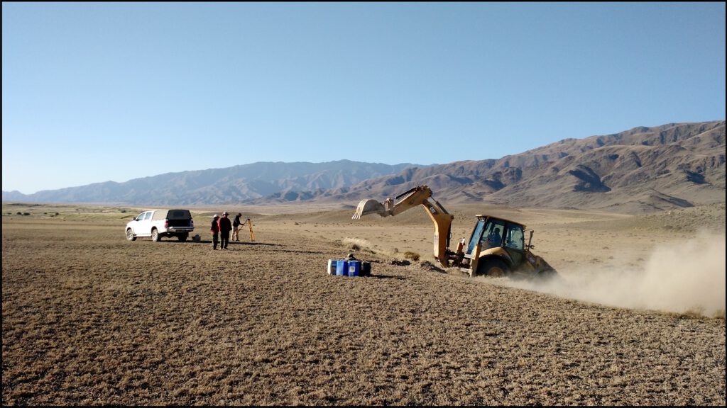 Photo of a vehicle and geologists standing on an arid plain with mountains in the background. In the foreground an excavator digs into a step in the plain, where an active fault has created a scarp